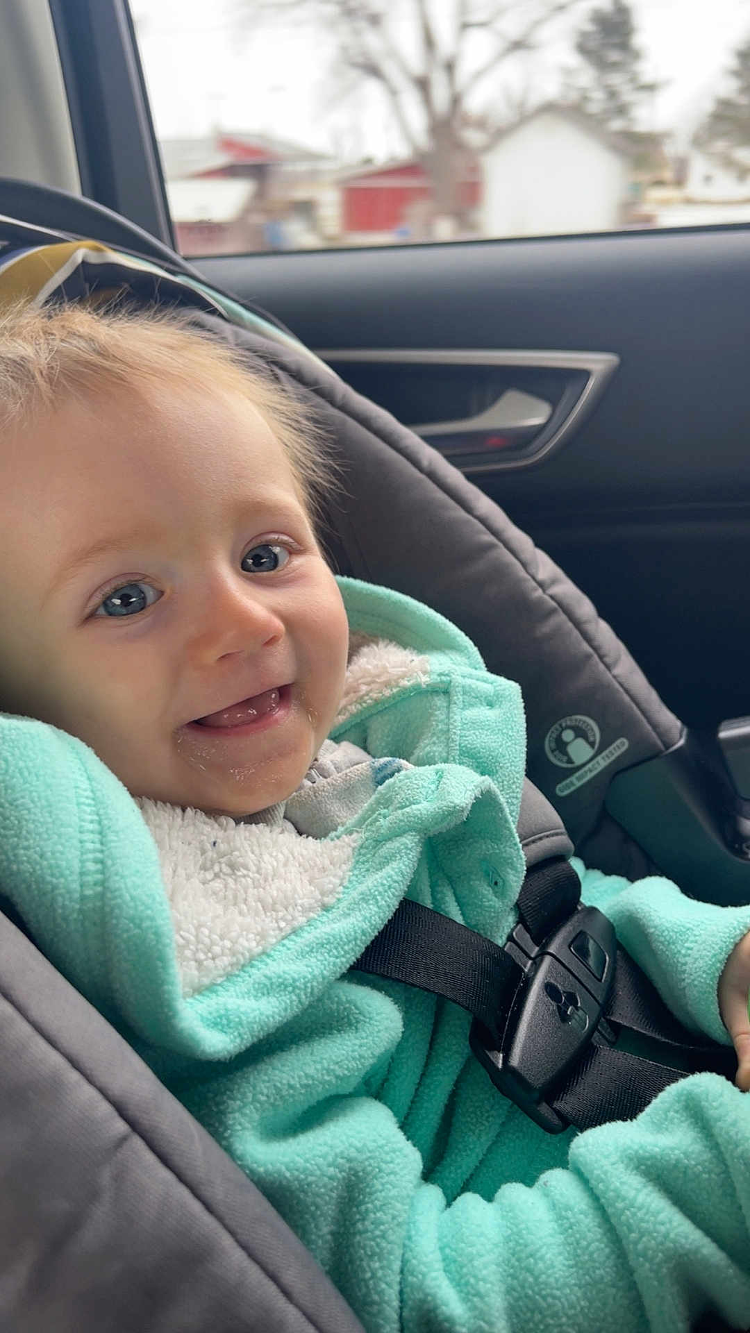 Melody is registered to the contest to win money with this photo: child, toddler, baby, car_seat, seatbelt, smile, happy, blue_eyes, mint_fleece, hooded_jacket, fleece, window, car_interior, outdoor, house, tree, drool, teeth, closeup, portrait