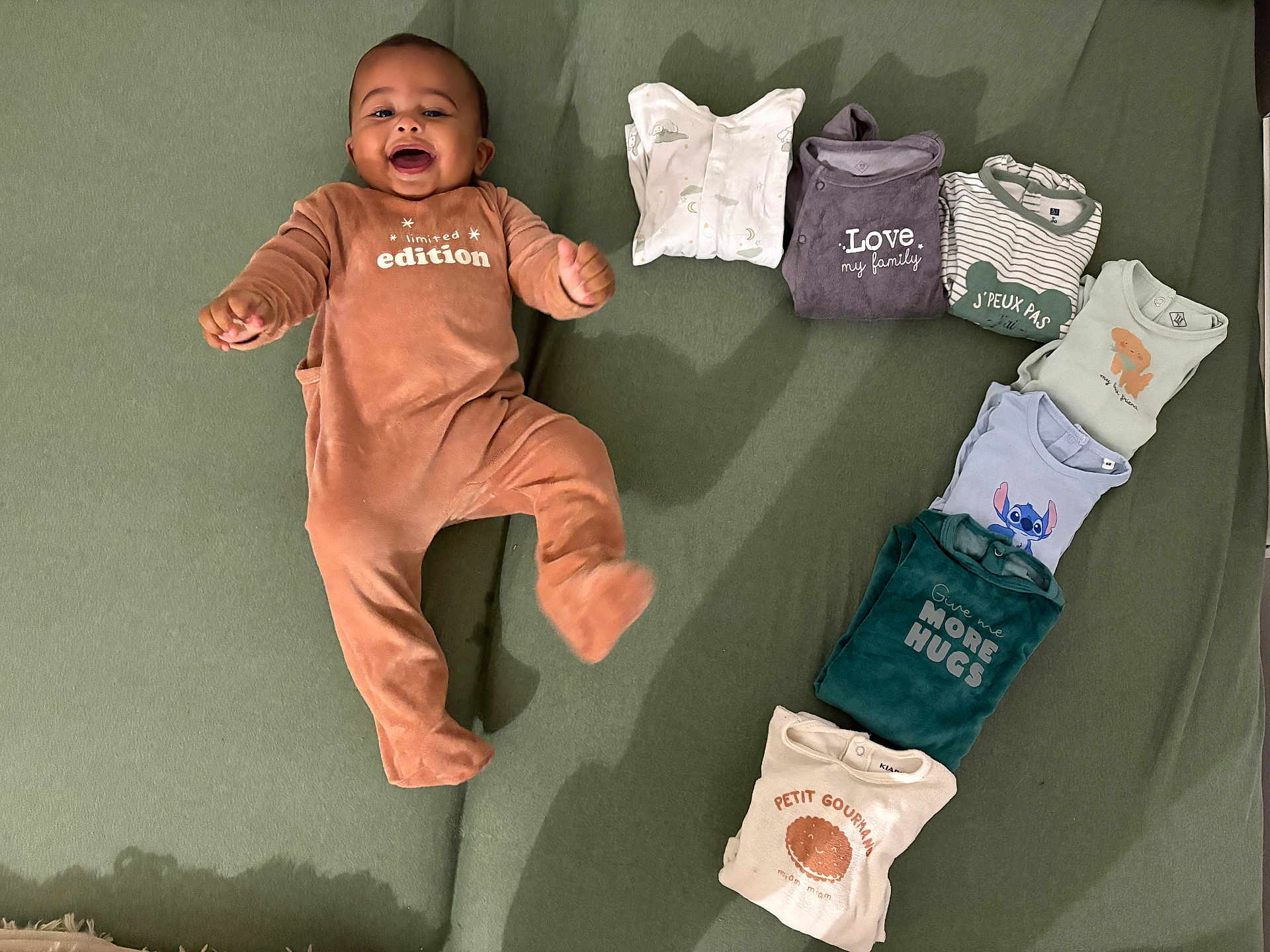 Lyvio a rejoint le concours — aidez-le/la à gagner de superbes lots ! baby, infant, onesie, clothing, folded_clothes, green_blanket, smiling, happy, lying_down, cute, child, indoors, fabric, soft_texture, cozy, playful, neatly_arranged, baby_fashion, person, text_on_clothing