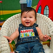 Manuel is registered to the contest to win money with this photo: baby, child, smiling, denim_overalls, wicker_chair, indoor, colorful_background, cartoon_characters, happy, cute, portrait, playful, clothing, person, infant, seated, furniture, bright_colors, expression, young_child