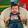 baby, child, smiling, denim_overalls, wicker_chair, indoor, colorful_background, cartoon_characters, happy, cute, portrait, playful, clothing, person, infant, seated, furniture, bright_colors, expression, young_child