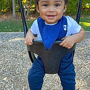 Mateo is registered to the contest to win money with this photo: toddler, child, swing, playground, smiling, curly_hair, blue_bib, white_shirt, blue_pants, socks, outdoor, trees, autumn, woodchips, daylight, happy, person, park, nature, play