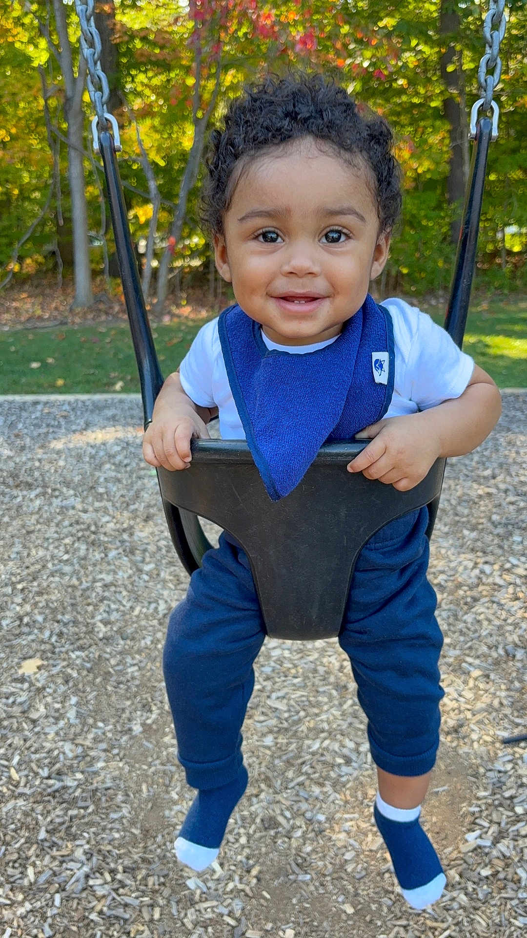 Mateo is registered to the contest to win money with this photo: toddler, child, swing, playground, smiling, curly_hair, blue_bib, white_shirt, blue_pants, socks, outdoor, trees, autumn, woodchips, daylight, happy, person, park, nature, play