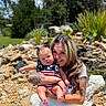 Rayleigh is registered to the contest to win money with this photo: woman, baby, tattoos, outdoor, garden, rocks, greenery, smiling, holding, child, summer, sunny, grass, plants, inflatable_slide, casual_clothing, portrait, family, happy, nature