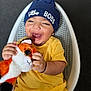 baby, child, smiling, hat, blue_hat, yellow_clothing, plush_toy, fox_toy, happy, laughing, indoor, seated, basket_chair, cute, infant, playful, person, toy, headwear, young_child