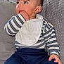 baby, bib, casual_clothing, child, comfort, couch, curious_look, cute, face, furniture, hand_in_mouth, indoor, infant, navy_pants, portrait, seated, soft_texture, striped_shirt, white_sock, young_child