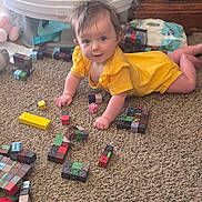 Rosalie is registered to the contest to win money with this photo: baby, child, crawling, yellow_outfit, smiling, portrait, indoor, carpet, building_blocks, toys, playroom, activity_center, infant, happy, face, brown_hair, sitting, floor, home, colorful