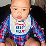 baby, child, infant, plaid_shirt, bib, sitting, cute, portrait, indoor, person, young_child, face, hand, legs, hair, clothing, expression, curious, red_heart, small_ponytail