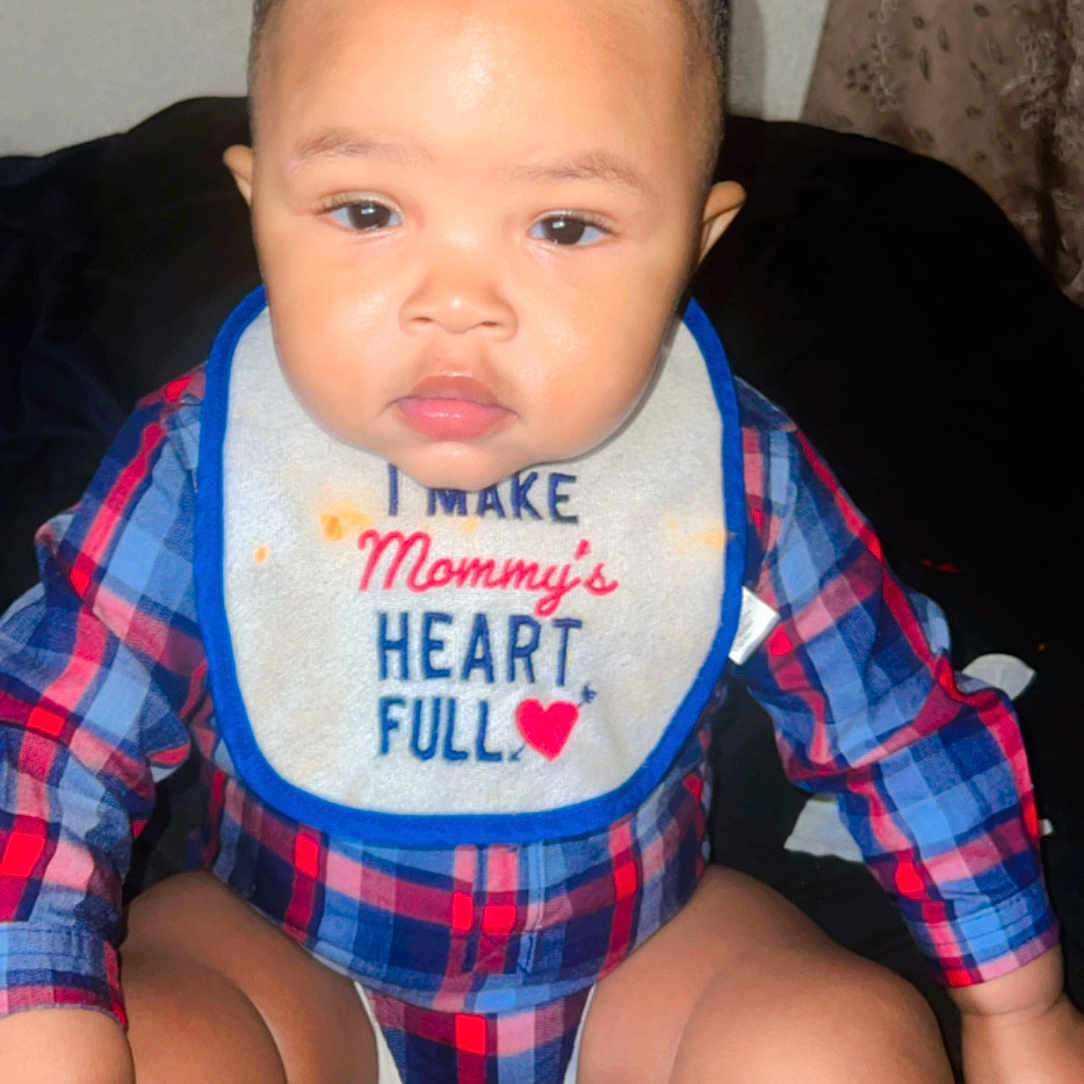 Antonio is registered to the contest to win money with this photo: baby, bib, child, clothing, curious, cute, expression, face, hair, hand, indoor, infant, legs, person, plaid_shirt, portrait, red_heart, sitting, small_ponytail, young_child