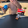 child, toddler, curly_hair, shirtless, outdoor, sunlight, portrait, face, eyes, hand, eating, snack, toy, blue_toy, red_wagon, playground, patio, table, wooden_deck, smiling