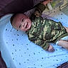 baby, infant, child, smiling, camouflage, onesie, bassinet, crib, blanket, bottle, nursery, sleeping_area, star_pattern, moon_pattern, lying_down, portrait, happy, indoor, face, infant_bed