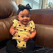 Emelia is registered to the contest to win money with this photo: baby, child, smiling, headband, bow, yellow_outfit, black_hearts, sitting, couch, leather, window, curtains, happy, indoors, person, infant, cute, portrait, black_tights, furniture