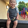 toddler, child, smiling, overalls, monkey_patch, sandals, wooden_railing, playground, play_structure, trees, outdoor, daylight, happy, person, short_sleeves, grass, blue_sky, wood_chips, fence, casual_clothing