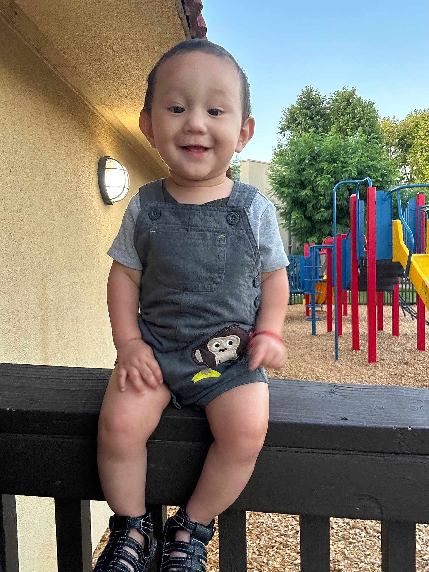 Jaziel joined the competition — help win amazing prizes! toddler, child, smiling, overalls, monkey_patch, sandals, wooden_railing, playground, play_structure, trees, outdoor, daylight, happy, person, short_sleeves, grass, blue_sky, wood_chips, fence, casual_clothing