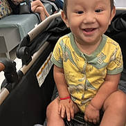 Jaziel is registered to the contest to win money with this photo: toddler, child, smiling, yellow_clothing, dinosaur_print, stroller, indoor, waiting_room, people, bench, shoes, happy, baby, casual, people_sitting, person, footwear, bracelet, blurred_background, hospital