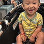 toddler, child, smiling, yellow_clothing, dinosaur_print, stroller, indoor, waiting_room, people, bench, shoes, happy, baby, casual, people_sitting, person, footwear, bracelet, blurred_background, hospital