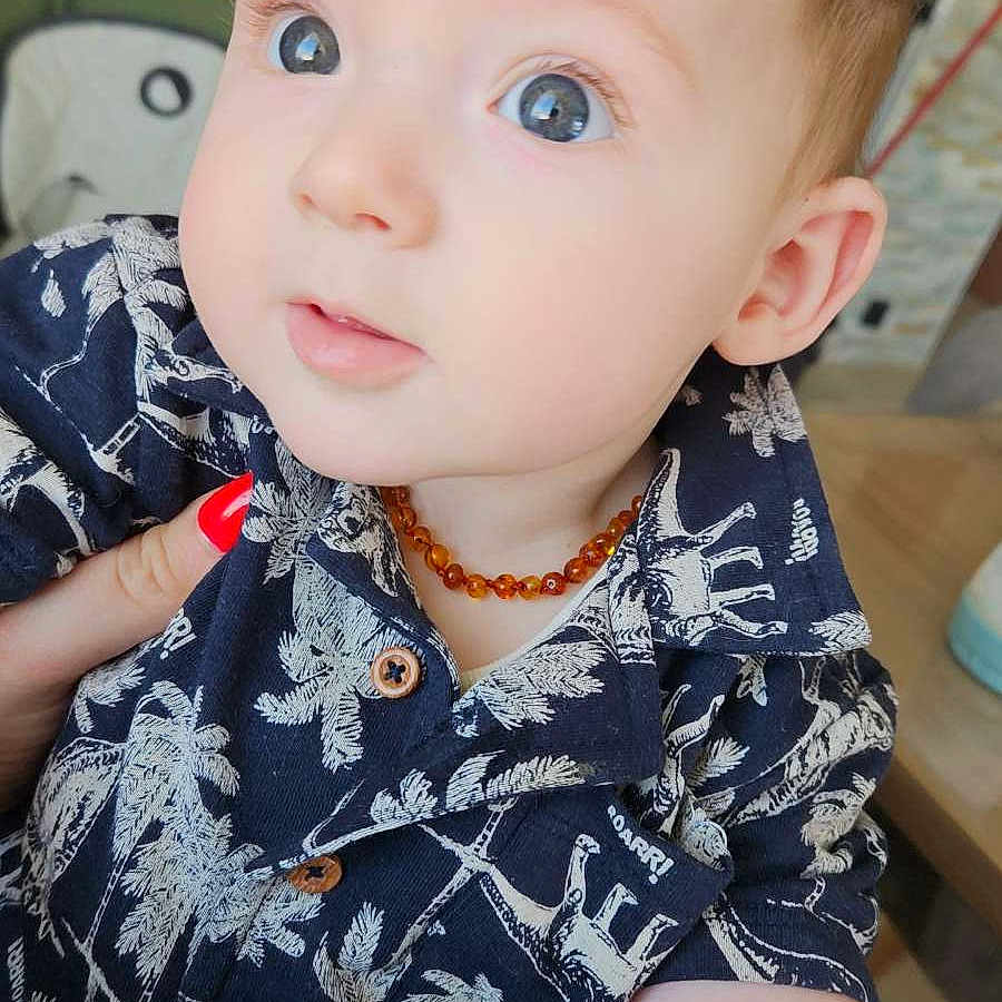 Gabriel participe au concours pour gagner de l'argent avec cette photo : amber_necklace, baby, blue_eyes, child, close_up, clothing, curious_look, cute, dinosaur_print, face, hand, holding, indoor, infant, navy_shirt, palm_tree_print, person, portrait, red_nail_polish, young_child