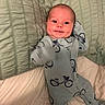 baby, bed, bicycle_pattern, blanket, blue_clothing, child, cute, face, happy, head, home, indoor, infant, lying_down, onesie, person, quilt, sleepwear, smile, soft_lighting