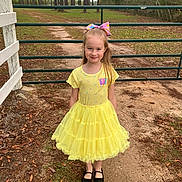 Venus is registered to the contest to win money with this photo: child, girl, yellow_dress, bow, outdoor, gate, path, trees, grass, smile, portrait, nature, happy, footwear, black_shoes, dirt_path, greenery, playful, casual_clothing, daylight