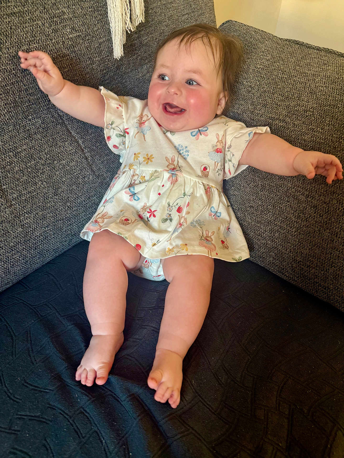 Daisy is registered to the contest to win money with this photo: baby, child, smiling, happy, arms_outstretched, dress, bunny_pattern, couch, seated, indoor, rosy_cheeks, barefoot, infant, cute, plush, portrait, person, clothing, furniture, home