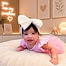 baby, child, infant, smiling, crawling, pink_dress, large_bow, headband, nursery, bed, blanket, stuffed_bunny, lamp, wall_art, heart, indoor, portrait, happy, decor, warm_lighting