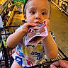 toddler, child, shopping_cart, grocery_store, snack_pouch, food, person, cute, baby, clothing, hand, retail, aisle, sitting, expression, face, supermarket, consumer_goods, colorful, young