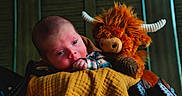 Neizan is registered to the contest to win money with this photo: baby, infant, child, plush_toy, monster_toy, horns, blanket, quilt, portrait, indoors, lying_down, wide_eyes, face, hand, clothing, warm_lighting, cozy, close_up, photography, nursery
