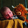 baby, infant, child, plush_toy, monster_toy, horns, blanket, quilt, portrait, indoors, lying_down, wide_eyes, face, hand, clothing, warm_lighting, cozy, close_up, photography, nursery