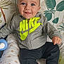 baby, black_pants, blue_eyes, child, cushion, cute, grey_clothing, happy, hoodie, indoor, infant, leaf_pattern, nike, patterned_cushion, portrait, seated, smiling, socks, tongue_out, wallpaper