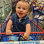 aisle, baby, black_bag, blue_shirt, child, clothing, face, grocery_store, hand, happy, indoor, luggage, people, person, retail, shopping, shopping_cart, smiling, store, supermarket