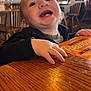 toddler, child, smiling, happy, restaurant, menu, wooden_table, indoor, person, clothing, face, hand, furniture, chair, cozy, casual, portrait, light, background, messy_face
