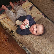 Noah is registered to the contest to win money with this photo: baby, bottle, pillow, rug, floor, child, infant, lying_down, home_interior, wood_floor, blanket, plastic_container, toy_storage, casual_clothing, curious_expression, hands, feet, blue_shirt, gray_pants, comfortable