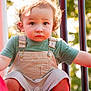 toddler, child, curly_hair, overalls, green_shirt, playground, slide, outdoor, daylight, face, eyes, expression, legs, shoes, candid, person, summer, park, young_child, nature