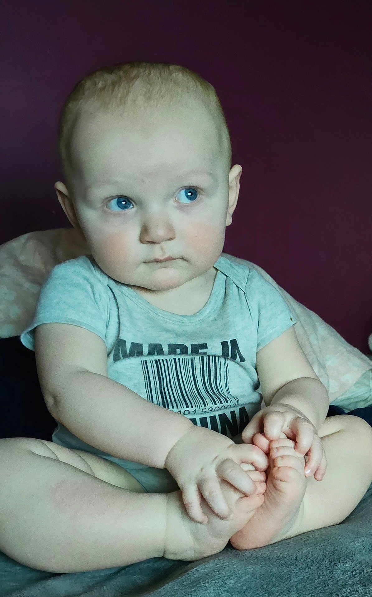 Casanova is registered to the contest to win money with this photo: baby, child, blue_eyes, sitting, onesie, barcode_shirt, barefoot, feet, hands, portrait, indoor, plum_wall, blanket, bed, cute, expression, head, short_hair, ears, skin