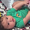 baby, child, infant, portrait, lying_down, bed, bedspread, pillow, plush_toy, green_shirt, yellow_toy, brown_hair, face, eyes, smiling, indoor, home, colorful, casual, photography