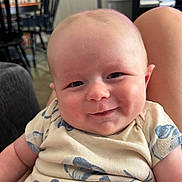 Bruin is registered to the contest to win money with this photo: baby, infant, child, smiling, portrait, close_up, face, eyes, indoor, home, seated, onesie, clothing, arms, head, happy, cute, dining_area, television, adult_arm