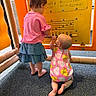 toddler, child, playground, musical_panel, yellow_panel, pink_clothing, blue_skirt, romper, barefoot, curly_hair, kneeling, standing, indoor_play_area, flooring, exploring, play, children, infant, interaction, colorful