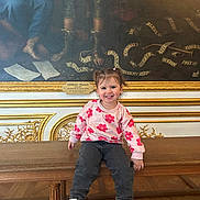 Umbelina participe au concours pour gagner de l'argent avec cette photo : child, toddler, smile, bench, painting, museum, indoor, ornate_frame, gold_trim, floral_sweater, jeans, sneakers, pigtails, hair_bows, wooden_bench, plaque, portrait_painting, happy, sitting, cute