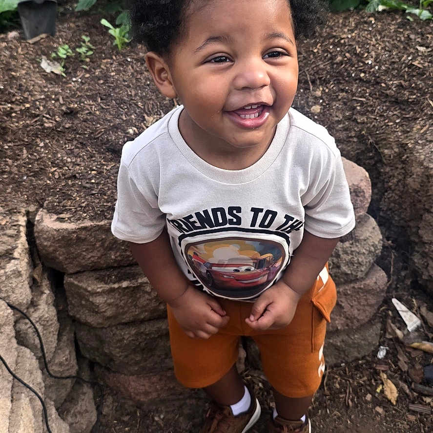 Taveon is registered to the contest to win money with this photo: toddler, child, smiling, curly_hair, outdoor, garden, plants, soil, stone, shoes, shorts, tshirt, happy, person, standing, nature, daylight, cute, young, playful