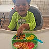 Taveon is registered to the contest to win money with this photo: toddler, child, high_chair, curly_hair, green_shirt, smiling, food, plate, fork, scrambled_eggs, fruit, pancakes, indoor, table, curtain, wooden_floor, happy, meal, baby, person