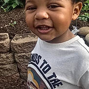 Taveon is registered to the contest to win money with this photo: child, toddler, smiling, outdoor, curly_hair, shirt, pants, stone_wall, garden, plants, greenery, standing, happy, person, casual_clothing, daylight, cute, portrait, young, nature