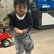 Kharina Kanistan participe au concours pour gagner de l'argent avec cette photo : child, toddler, curly_hair, indoor, floor, plaid_pants, black_top, white_sneakers, toy_car, television, plant, wood_floor, holding_object, furniture, living_room, person, young_child, casual_clothing, home