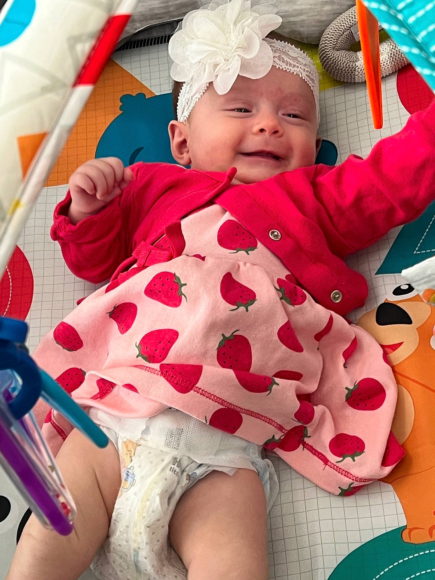 Penelope joined the competition — help win amazing prizes! baby, child, colorful, cute, diaper, dress, flower, happy, headband, indoor, infant, lying_down, pink_clothing, play_mat, reaching, red_cardigan, smiling, strawberry, toy, young_child