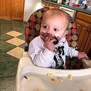 Louis joined the competition — help win amazing prizes! baby, high_chair, chocolate, messy, cookie, kitchen, wooden_cabinets, tiled_floor, child, food, face, hand, white_sweatshirt, colorful_chair, eating, happy, infant, indoor, person, snack
