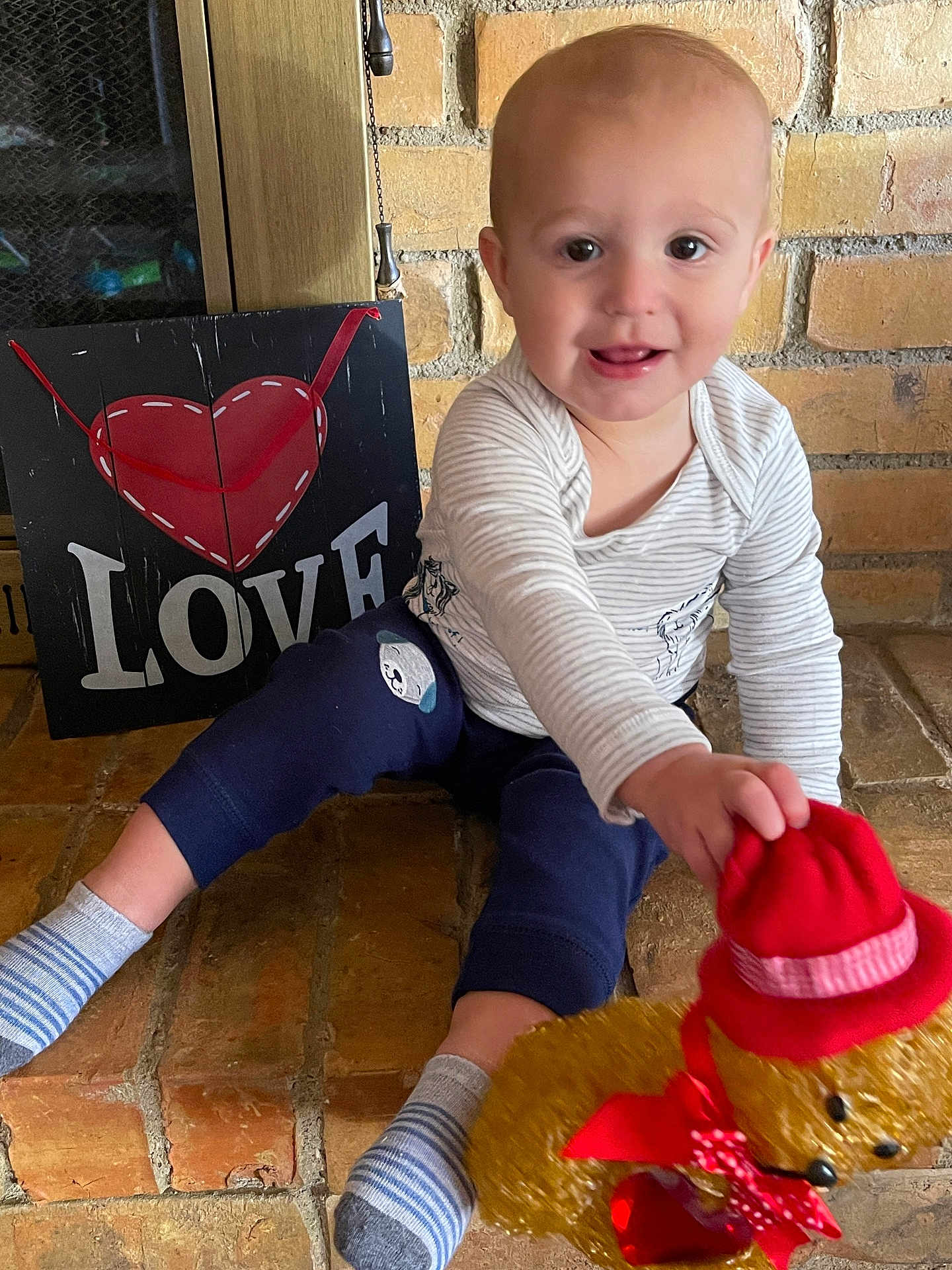 Louis joined the competition — help win amazing prizes! baby, child, teddy_bear, red_hat, red_scarf, brick_floor, love_sign, striped_shirt, blue_pants, socks, smiling, indoor, toy, cute, person, playful, happy, portrait, floor, decor