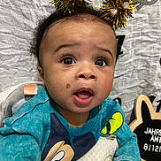 Jahreese is registered to the contest to win money with this photo: baby, child, face, expression, headband, tinsel, sweater, mickey_mouse, high_chair, hand, background, sign, indoors, cute, surprised, wide_eyes, open_mouth, skin, person, portrait