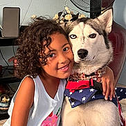 Bella is registered to the contest to win money with this photo: child, dog, siberian_husky, pet, indoor, curly_hair, hugs, smiling, heterochromia, face, closeup, white_wall, fan, flower_arrangement, red_harness, blue_harness, stars, casual_clothing, floor, affection