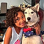 child, dog, siberian_husky, pet, indoor, curly_hair, hugs, smiling, heterochromia, face, closeup, white_wall, fan, flower_arrangement, red_harness, blue_harness, stars, casual_clothing, floor, affection