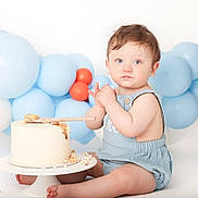 Dalton is registered to the contest to win money with this photo: baby, child, cake, birthday, balloons, blue, orange, romper, sitting, frosting, crumbs, wooden_spoon, celebration, cute, indoor, portrait, messy, smash_cake, light_background, happy