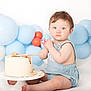 baby, child, cake, birthday, balloons, blue, orange, romper, sitting, frosting, crumbs, wooden_spoon, celebration, cute, indoor, portrait, messy, smash_cake, light_background, happy