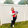 Sinclair is registered to the contest to win money with this photo: baby, casual_clothing, child, family, glasses, grass, headband, holding, infant, legs, nature, outdoor, parent, person, relaxed, smile, sneakers, tree, white_fence, woman